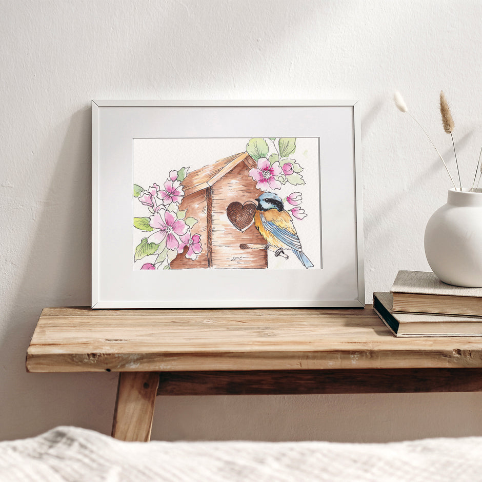 Framed artwork of a birdhouse with flowers on a wooden table.