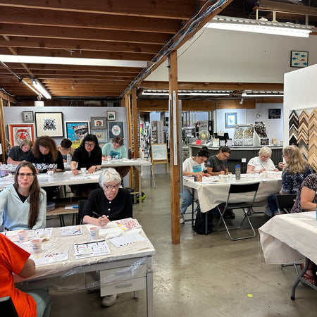 Art studio with people working at tables, surrounded by art supplies and framed artwork.