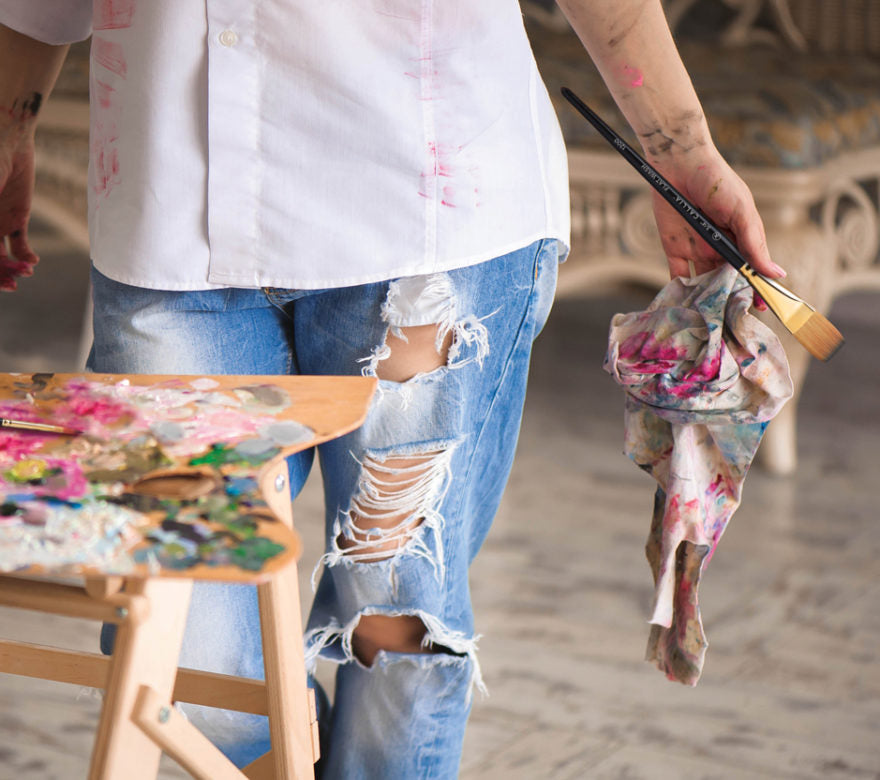 Person holding a paintbrush and a paint-splattered cloth, standing next to a stool with art supplies.